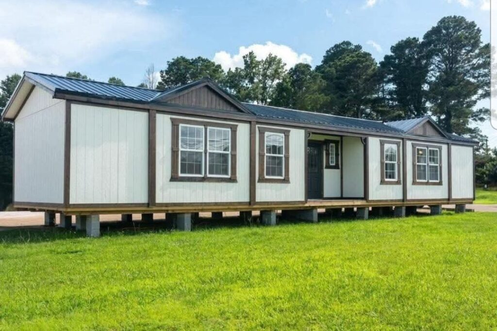 Single-story modular home with white siding, brown trim, and a black roof, set on raised blocks. Surrounded by lush grass and tall trees, under a clear sky.