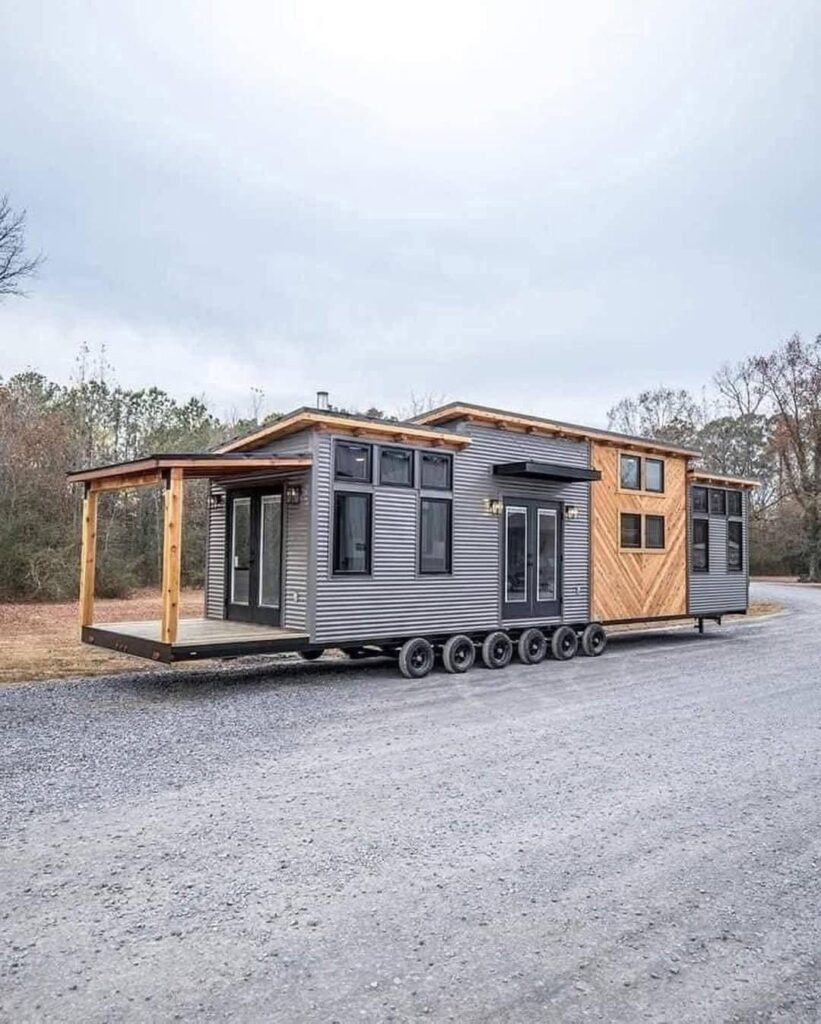 A modern tiny house on wheels with wood and metal siding. It features large windows, a deck, and is set on a gravel path surrounded by trees under a cloudy sky.