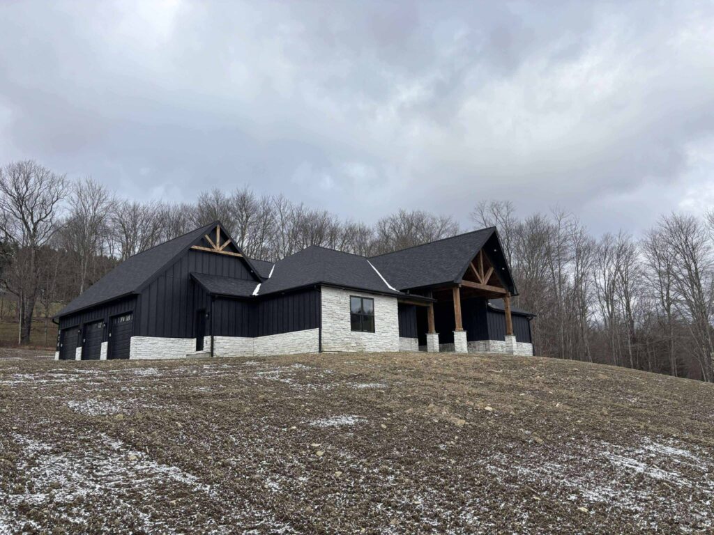 Modern black farmhouse with stone accents sits on a bare hillside under cloudy skies, surrounded by leafless winter trees, conveying a serene, rustic feel.