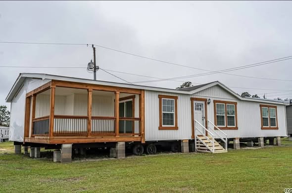 A white mobile home on blocks, featuring a wooden porch and small steps leading to the door, sits under a cloudy sky. The mood is calm and tranquil.