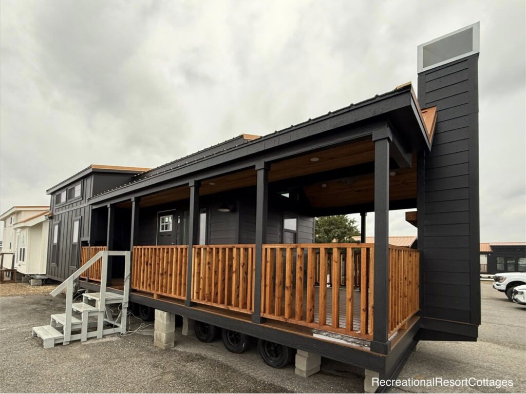 Black tiny house on wheels with a wooden porch railing and exterior staircase. The sky is overcast, creating a cozy and inviting atmosphere.