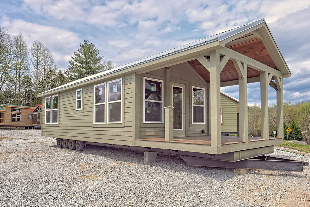 Small green mobile home on wheels with a wooden front porch. Set on a gravel lot with trees in the background under a partly cloudy sky.