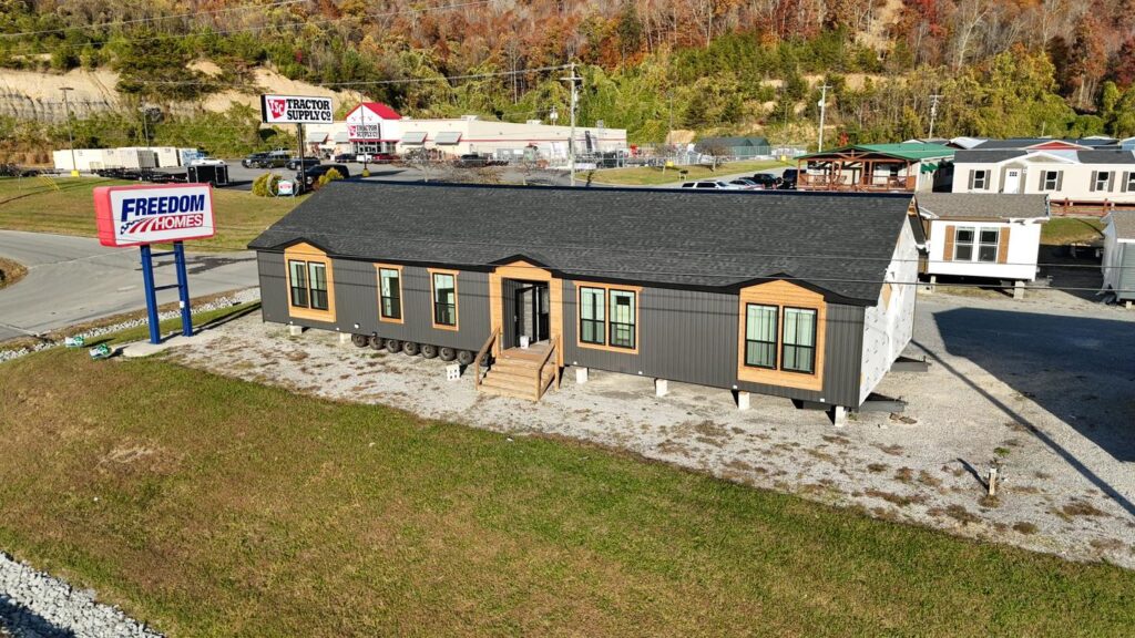 A large manufactured home with a dark roof sits on a gravel lot beside a "Freedom Homes" sign. Nearby is a Tractor Supply store and wooded hills.