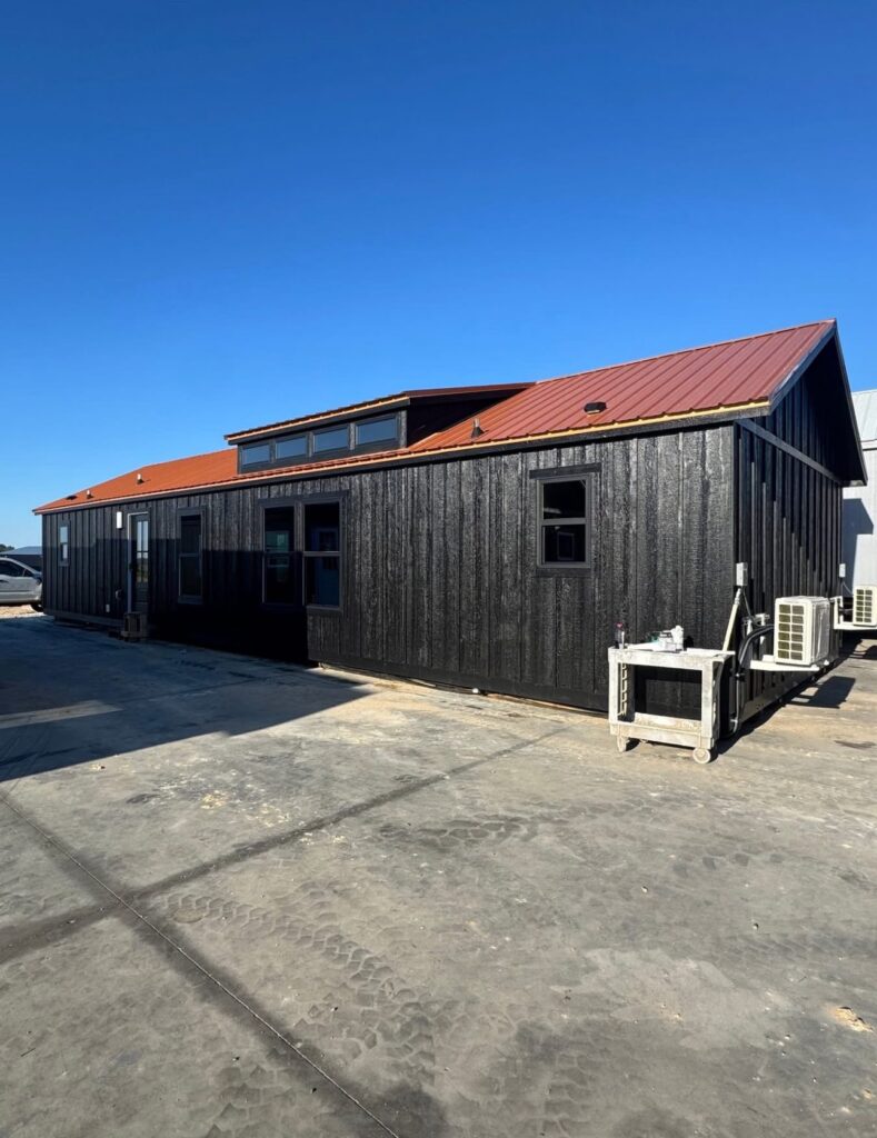 Modern tiny home with a red metal roof and black wooden siding under a clear blue sky. It sits on a wide concrete lot, conveying a minimalist vibe.