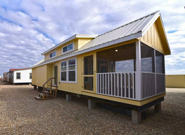 Yellow tiny home with a metal roof and screened porch, elevated on stilts. It sits on a gravel lot under a sky filled with scattered clouds.