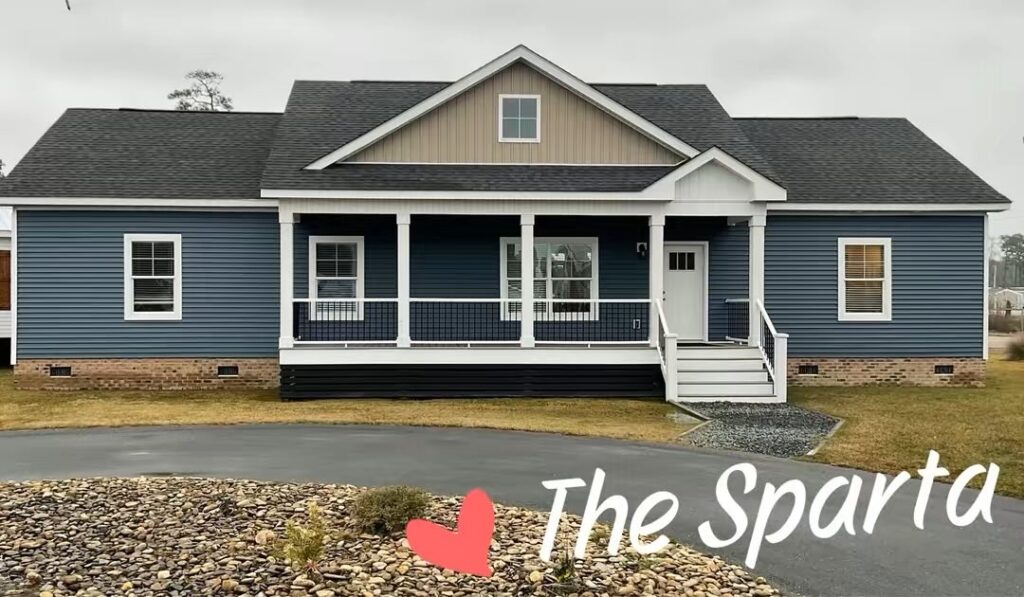 Blue, single-story house with a gabled roof and a front porch. The yard has a paved driveway and a stone garden. "The Sparta" is written with a heart symbol.