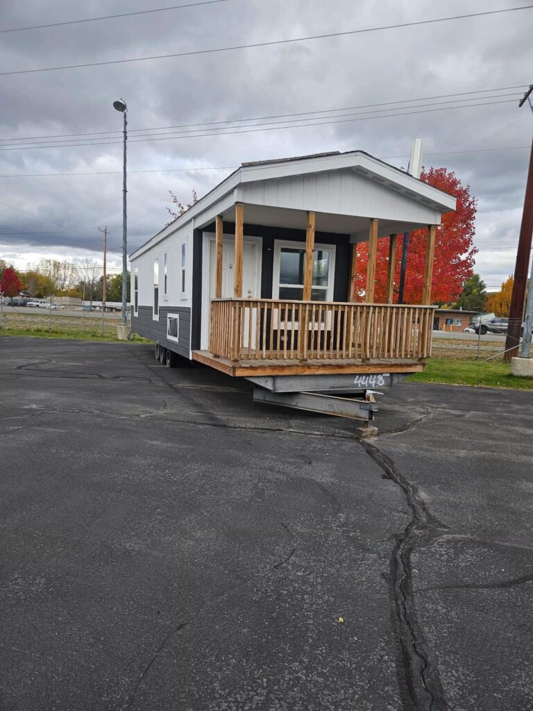 A small mobile home with a porch sits in a parking lot under overcast skies. The home is grey and white, with autumn trees in the background.